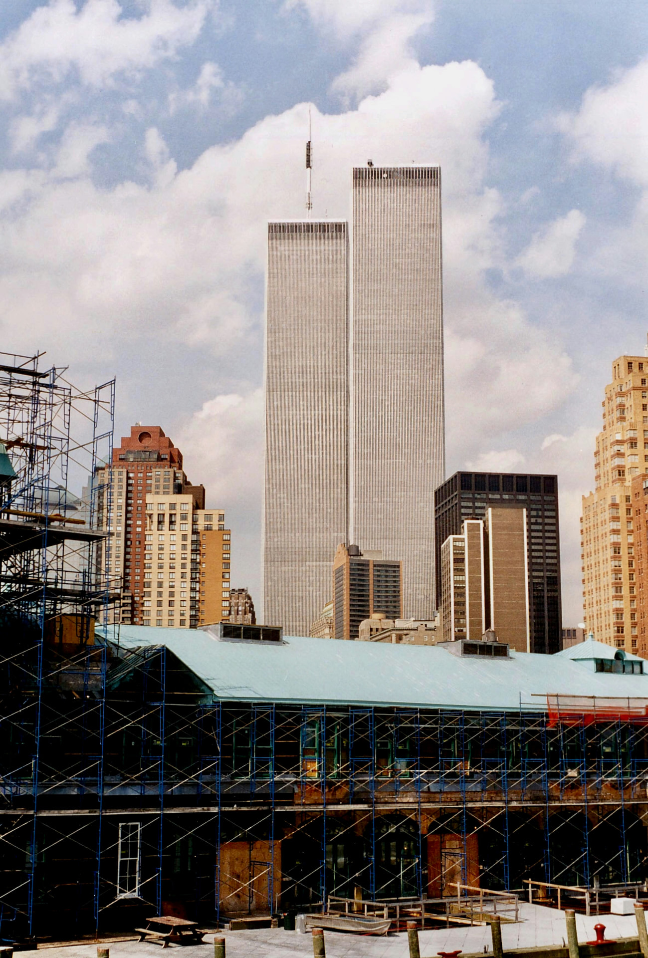 A daytime view of the World Trade Center Twin Towers in Lower Manhattan, rising behind a building covered in blue construction scaffolding under a cloudy sky
