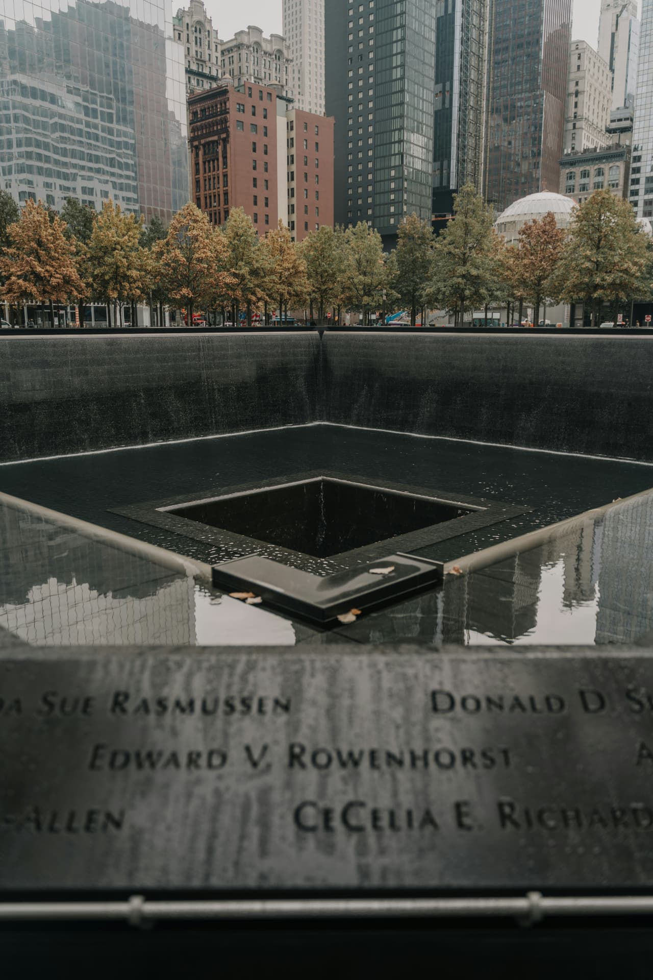 The 9/11 Memorial reflecting pool in Lower Manhattan, with victim names inscribed on the bronze parapet and surrounding skyscrapers reflected in the still water