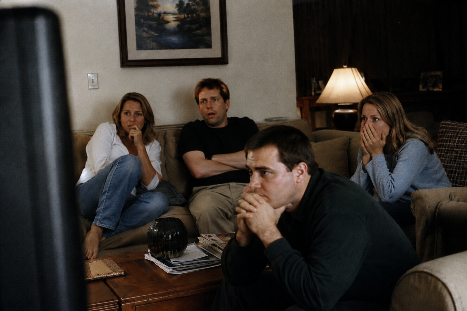 Four adults seated on a living room couch, watching a television in shock and distress